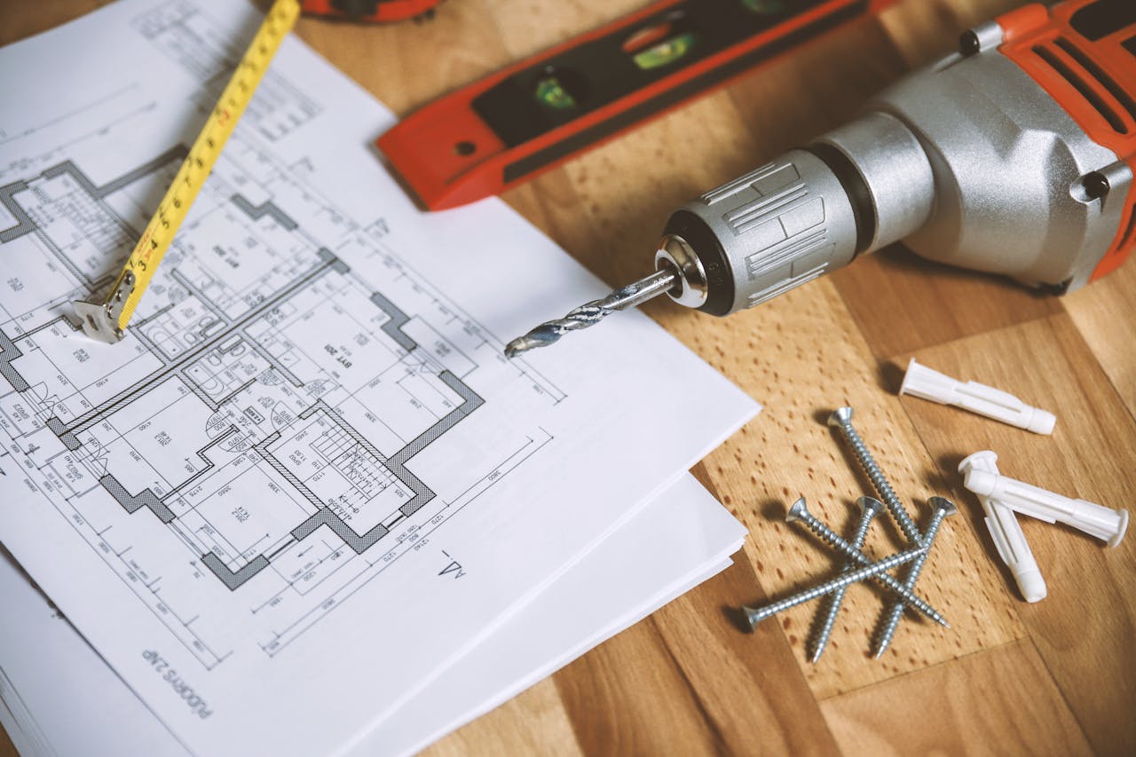 heros-img Close-up of construction blueprint with tools like drill and screws on a wooden table.